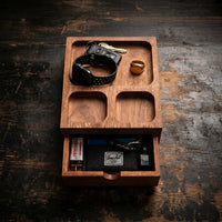 Thumbnail for A wooden organizer tray with a drawer, shown open on a dark rustic wooden table. The tray has three upper compartments holding a black smartwatch band, keys, and a small gold ring, and a lower drawer containing a black pouch with a Herschel label and other small accessories.

